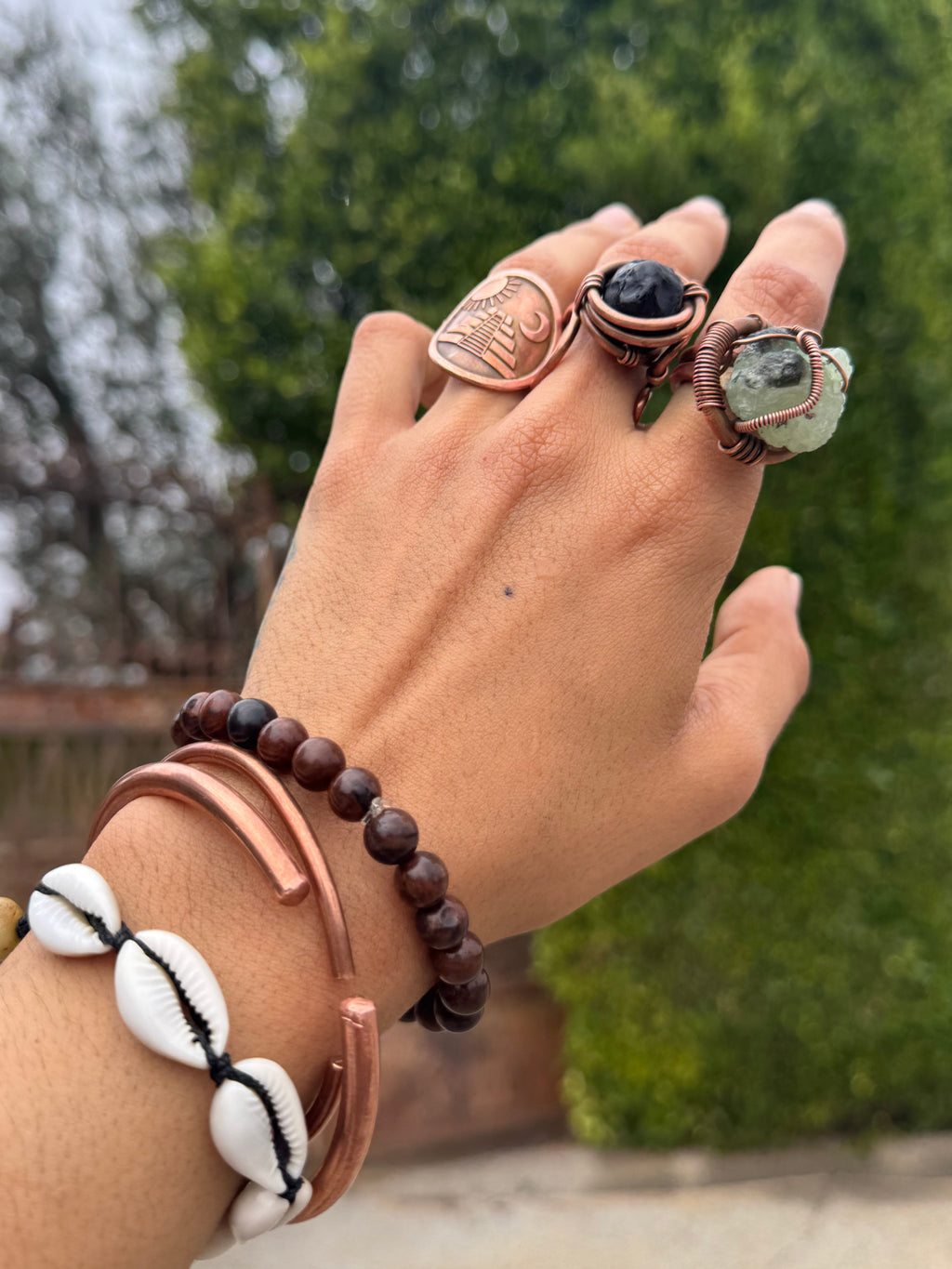 Hand wearing multiple bracelets and rings with a blurred natural background